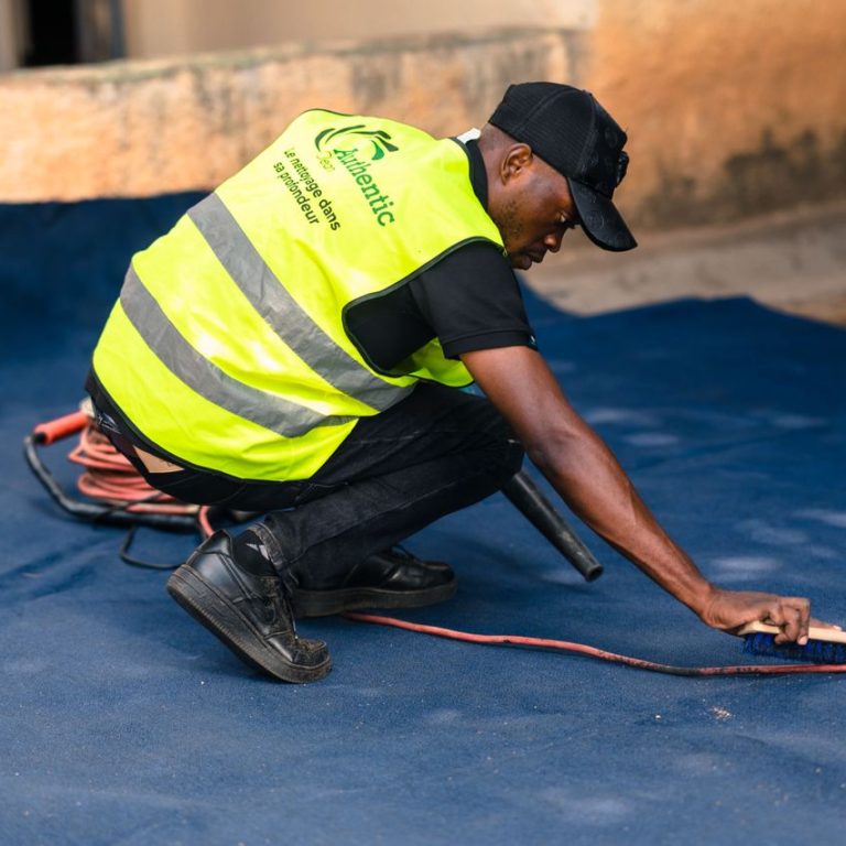 A worker dressed in a safety vest cleans a blue carpet outdoors with a brush.