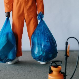 Sanitation worker in orange overalls holding blue trash bags indoors for waste management.
